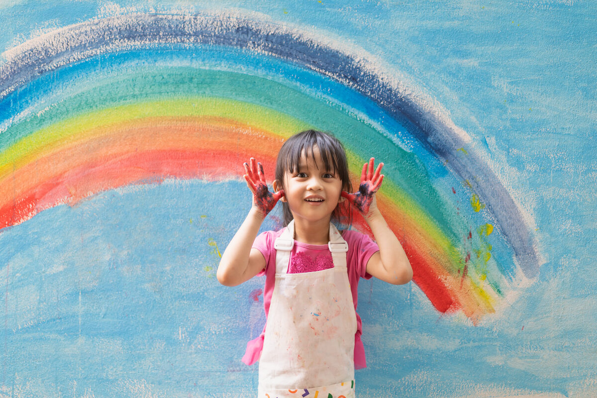 A child smiling proudly in front of a rainbow she painted, shared by her nanny